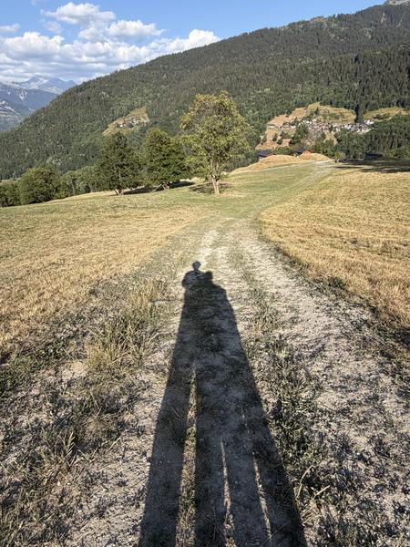This photo captures two long shadows stretching across a dry dirt path, set against a backdrop of lush green mountains under a bright blue sky at Chemin des Pommettes, Les Allues
